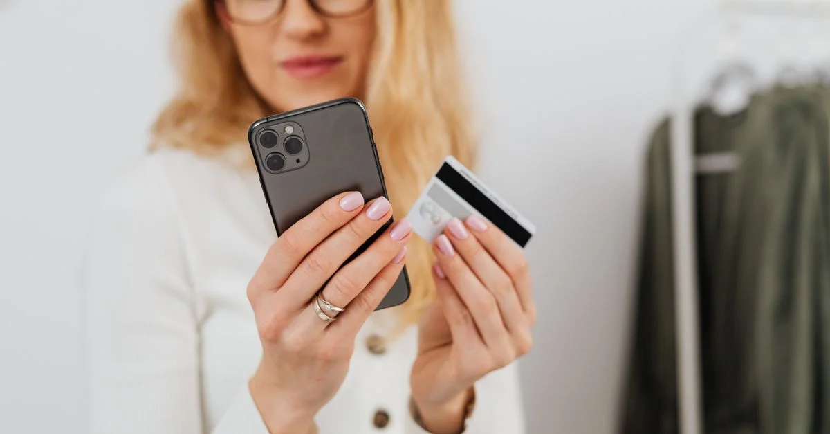 Une femme tenant un smartphone et une carte de crédit, participant à des achats en ligne à l'intérieur.