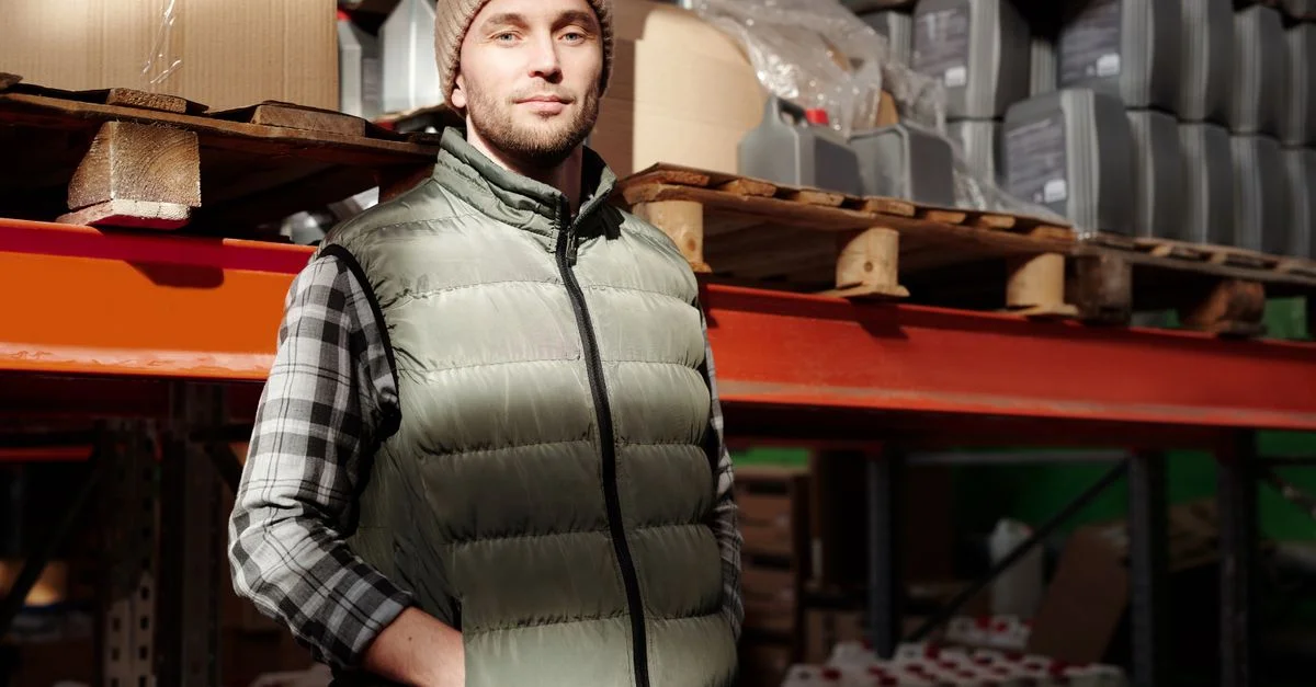 Un homme en gilet vert se tient confiantly dans un entrepôt, illuminé par la lumière naturelle.