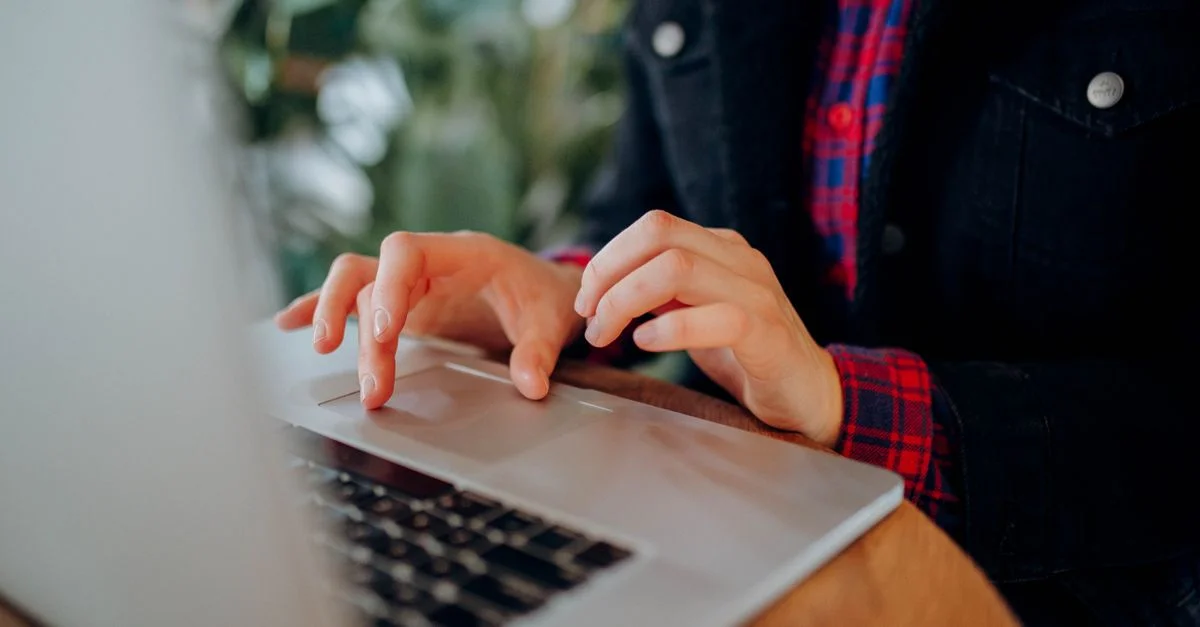Personne portant une chemise à carreaux tapant sur le clavier d'un ordinateur portable à l'intérieur, se concentrant sur les mains et l'appareil.