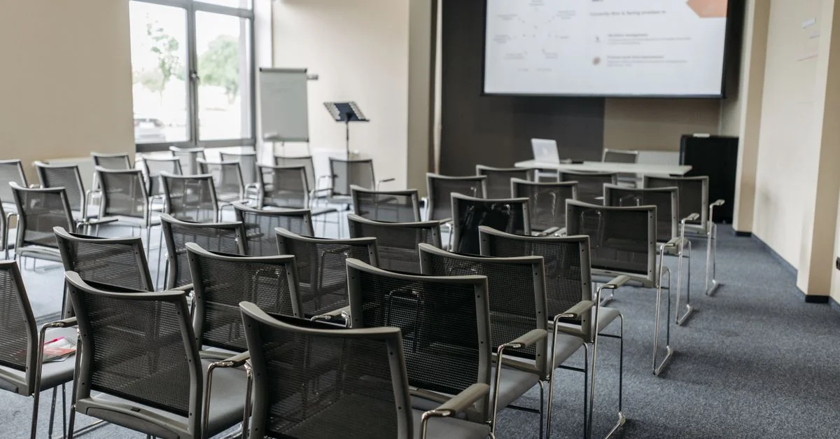 Salle de conférence spacieuse et vide avec des chaises disposées, un écran et une lumière naturelle pour des réunions professionnelles.