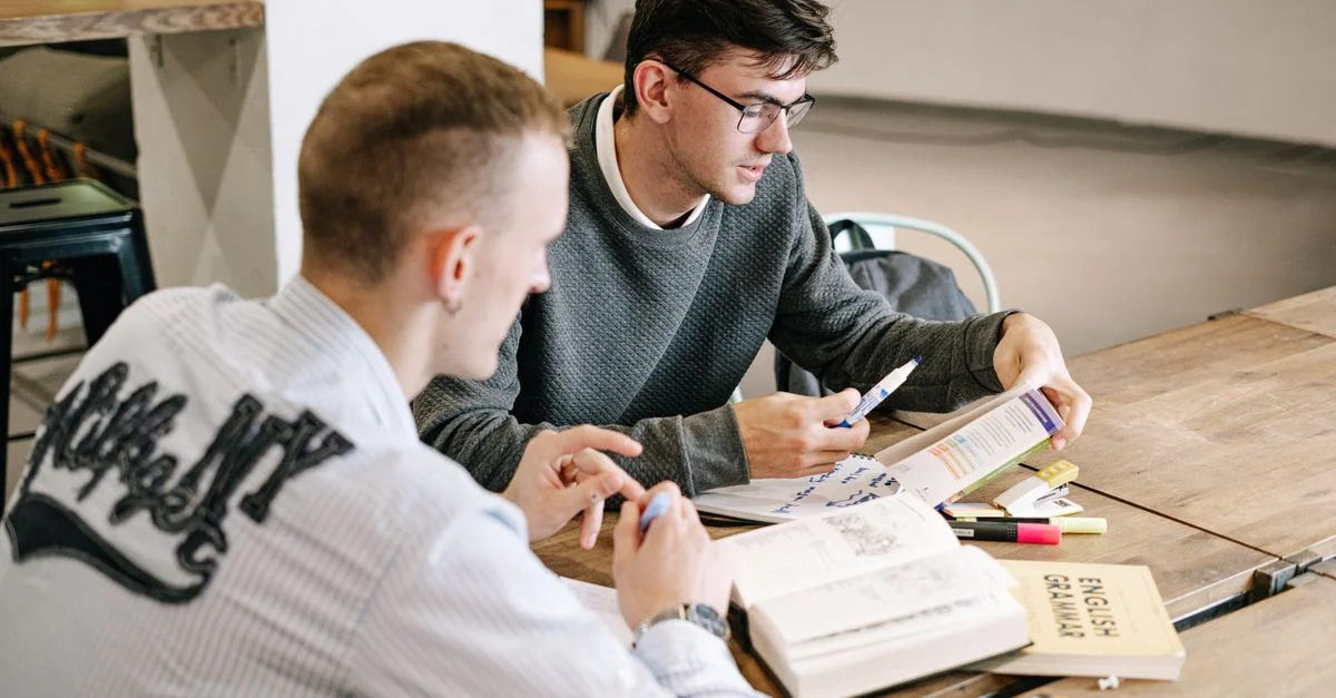 Deux jeunes hommes étudiant ensemble, concentrés sur des livres de grammaire anglaise dans un cadre intérieur moderne.