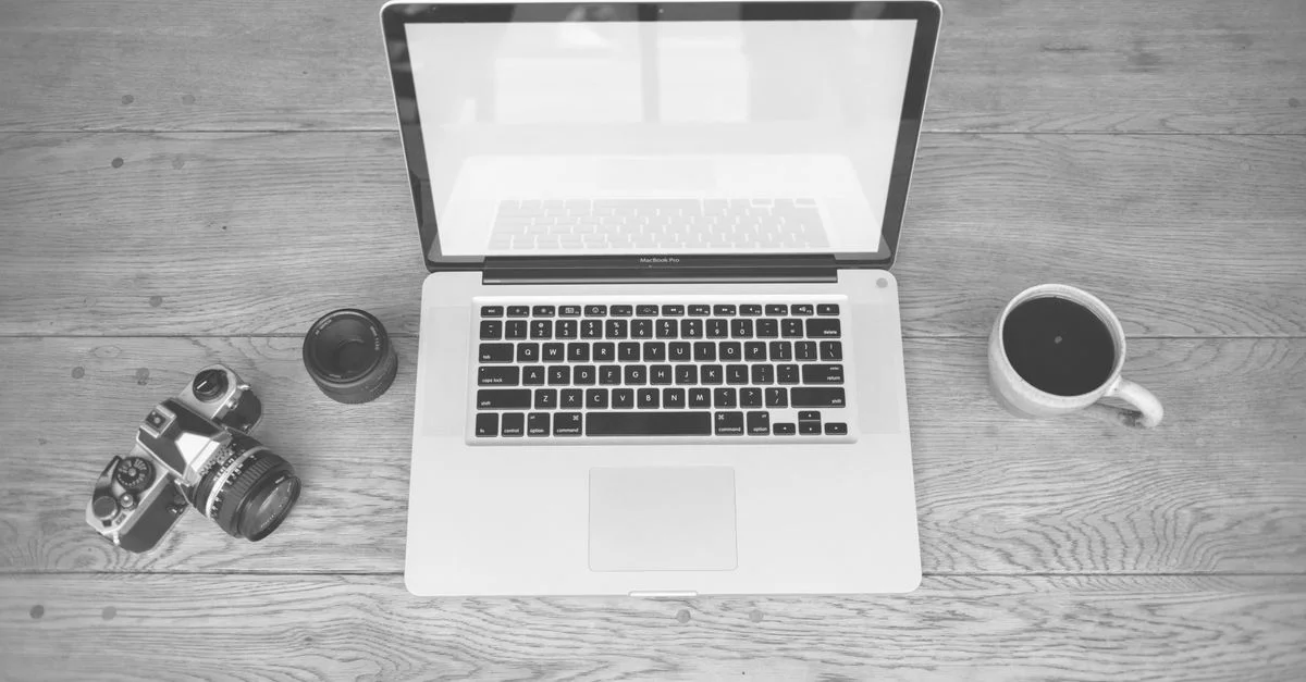 Image monochrome d'un ordinateur portable, d'une caméra, d'un objectif et d'une tasse de café sur un bureau en bois