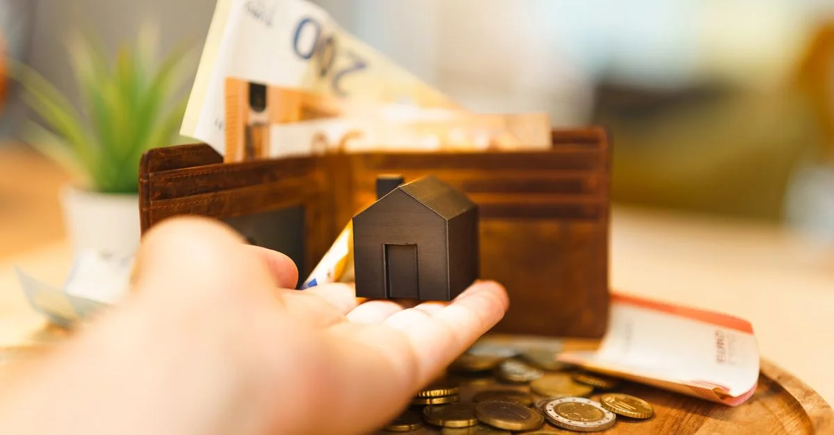 Une main tenant une petite maquette de maison avec des billets et des pièces en euros à proximité, illustrant l'investissement immobilier et la finance.