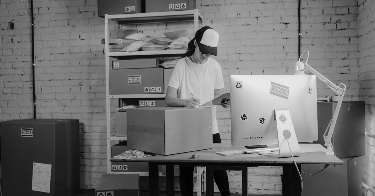 Une femme manipulant des colis dans un bureau monochrome, concentrée sur l'organisation des expéditions et de la logistique.
