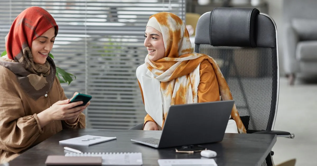 Deux femmes en hijab discutant de travail à un bureau moderne, mettant en avant la diversité et le travail d'équipe.