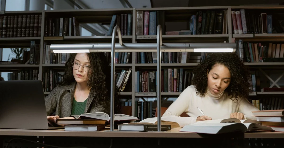Deux étudiantes avec des livres et des ordinateurs portables dans une bibliothèque moderne.