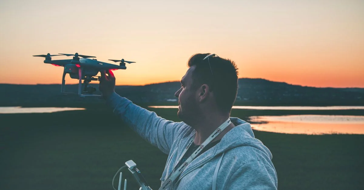 Un homme pilote un drone devant un magnifique coucher de soleil au bord d'un lac, mettant en valeur la technologie et la nature.