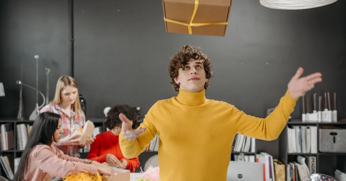 Un jeune homme en pull jaune lançant une boîte-cadeau dans un bureau créatif avec des collègues.