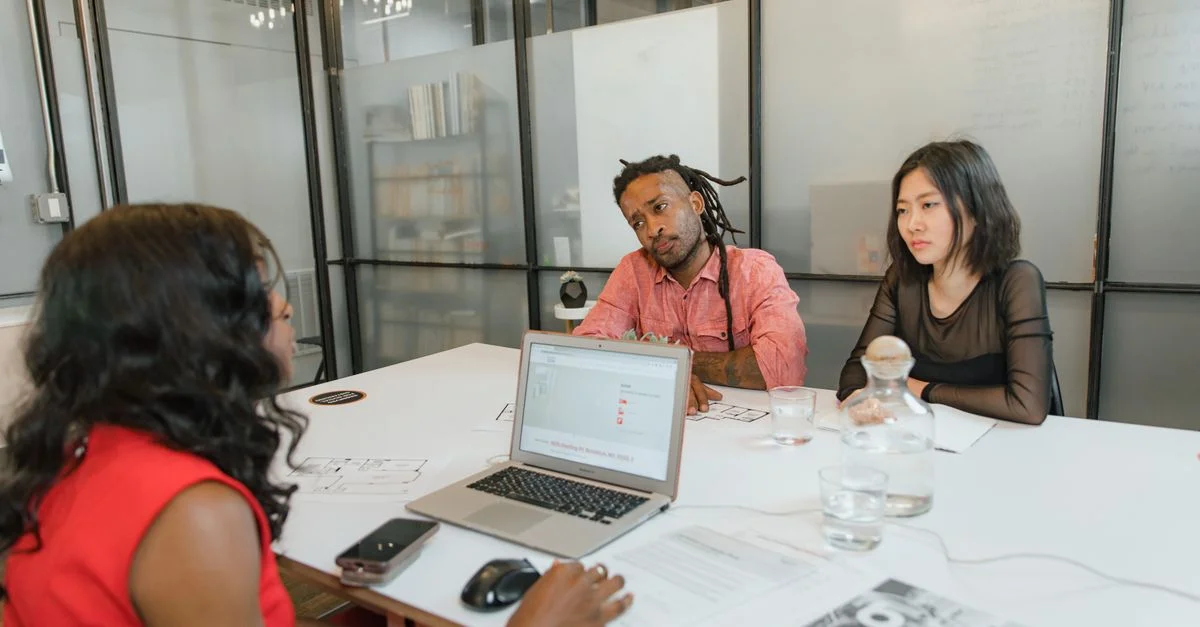 Trois professionnels participent à une réunion de bureau avec un ordinateur portable, des documents et des discussions.