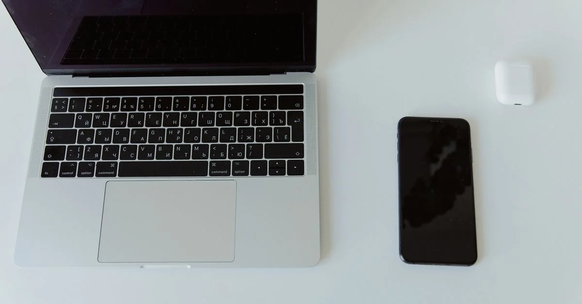 Espace de travail moderne et minimaliste comprenant un ordinateur portable, un smartphone et un étui d'écouteurs sans fil sur une table blanche.