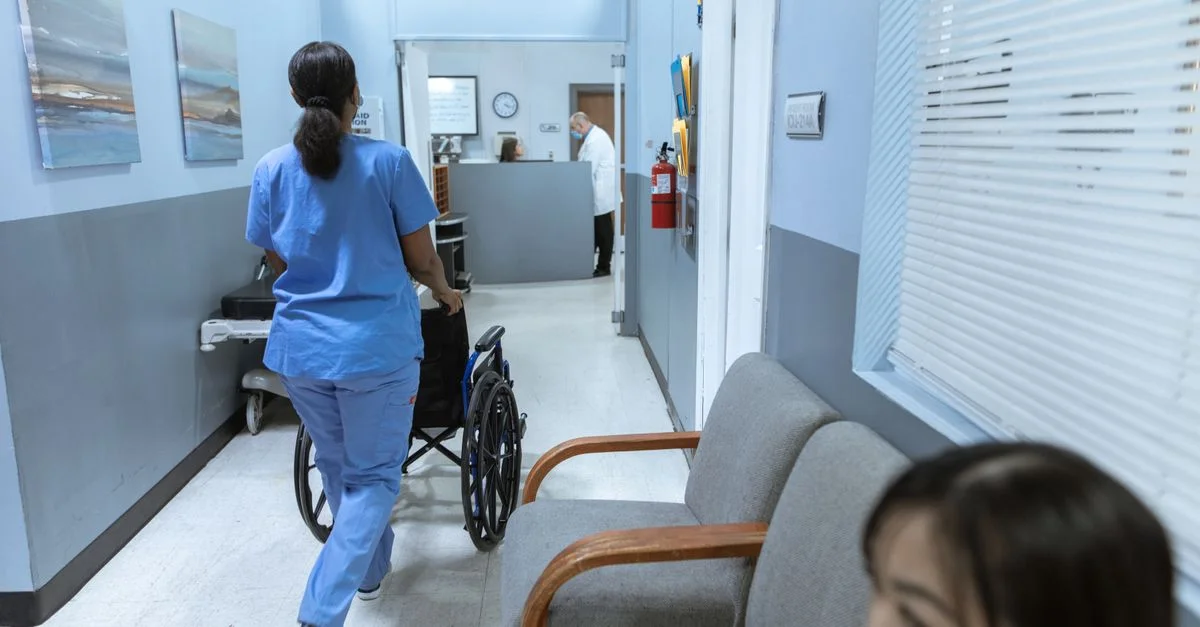 Une infirmière en tenue médicale pousse un fauteuil roulant à travers un couloir d'hôpital bien éclairé, s'occupant des soins aux patients.