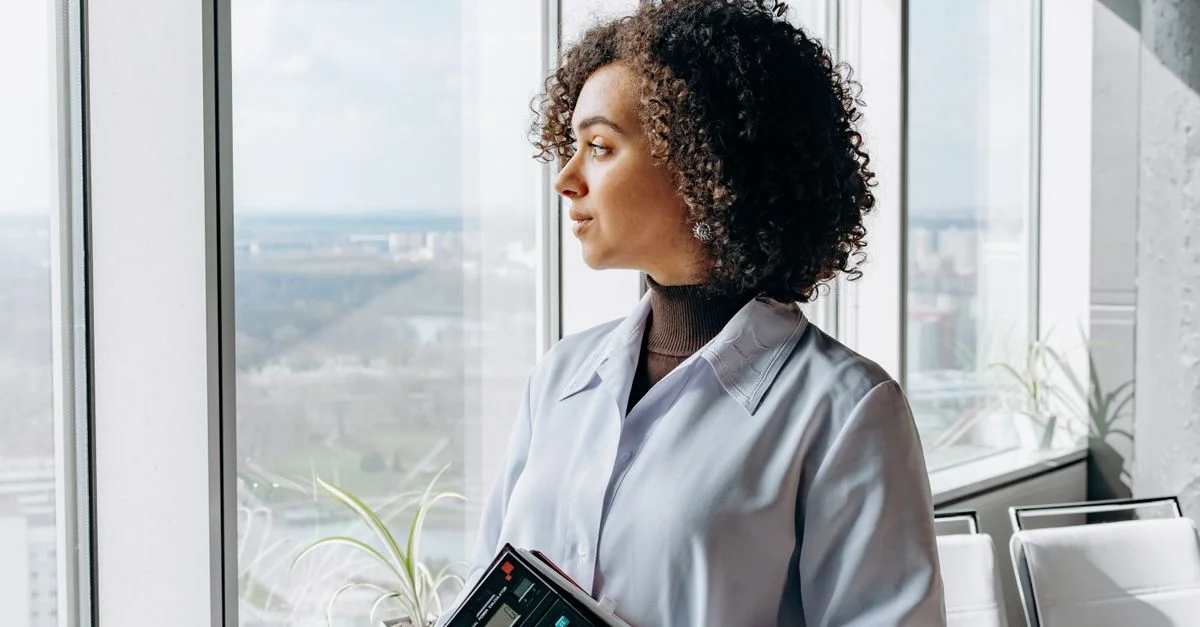 Femme en tenue de bureau tenant une calculatrice, regardant à travers une grande fenêtre, avec une vue sur la ville.