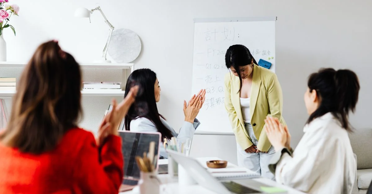 Des femmes asiatiques dans un bureau applaudissent une collègue après une présentation avec un tableau blanc en arrière-plan.