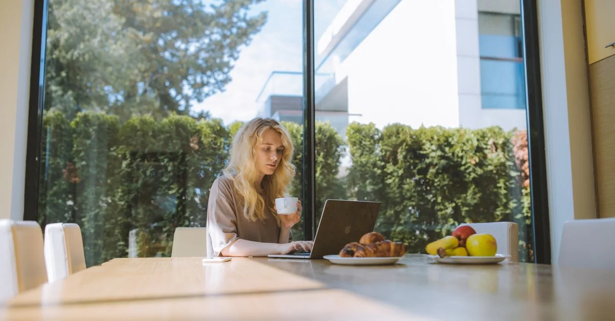 Femme blonde travaillant sur un ordinateur portable à la maison, appréciant un café avec des croissants