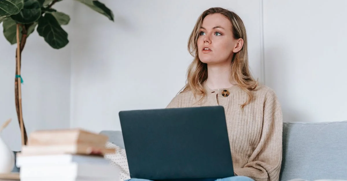 Jeune femme utilisant un ordinateur portable assise sur un canapé, travaillant à distance à la maison avec des livres à proximité.