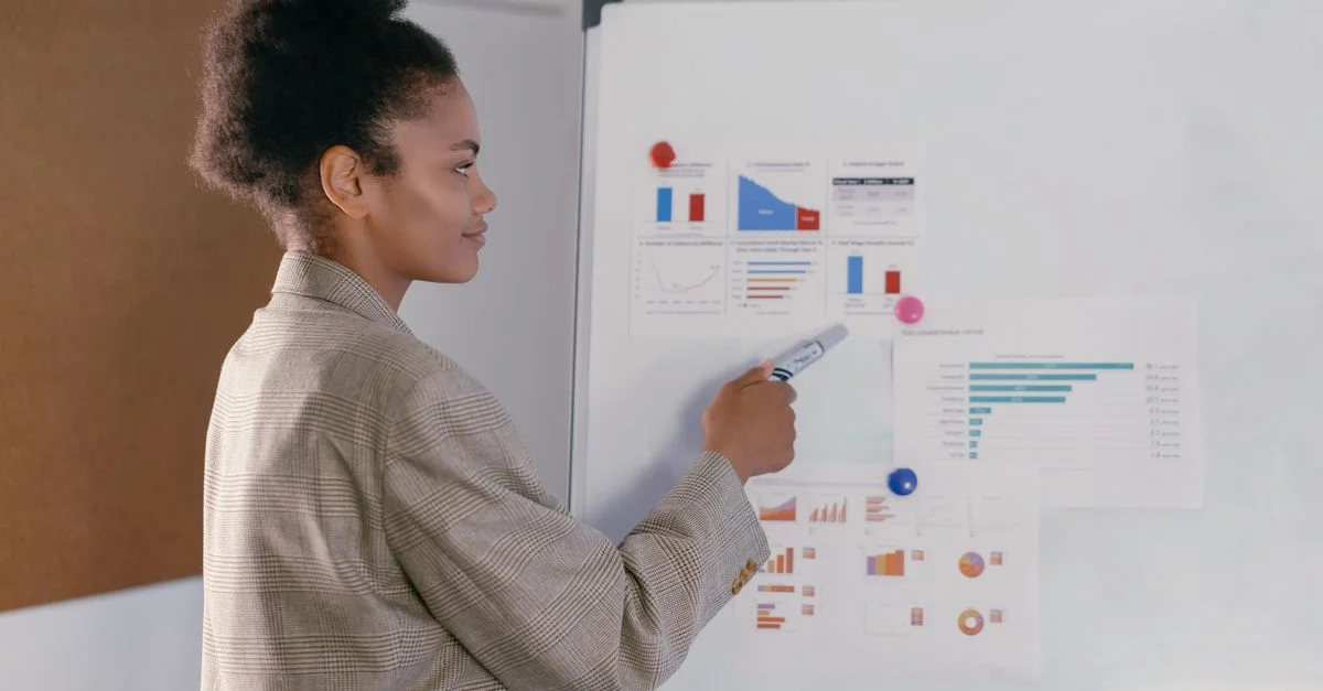 Femme professionnelle en blazer discute des graphiques financiers sur un tableau blanc lors d'une présentation.