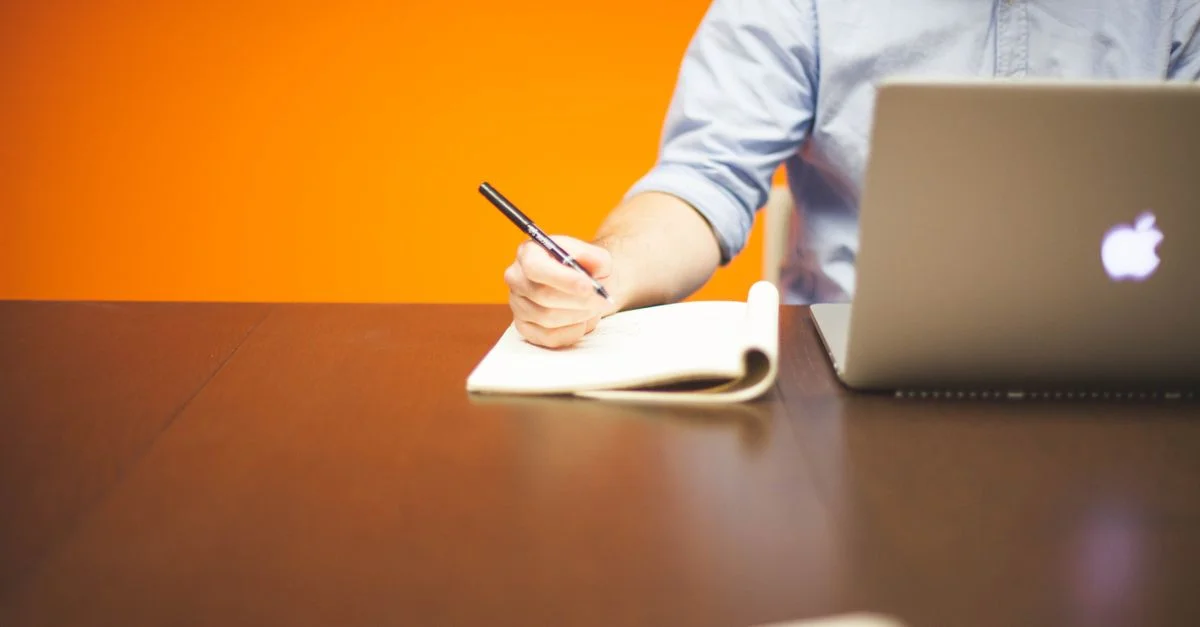 Un adulte prenant des notes dans un carnet à côté d'un ordinateur portable sur un bureau en bois avec un fond orange.
