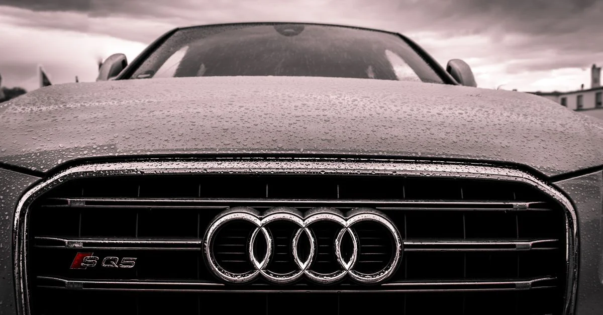 Gros plan sur une grille de voiture Audi couverte de pluie, mettant en avant le luxe et l'élégance sous des cieux nuageux.