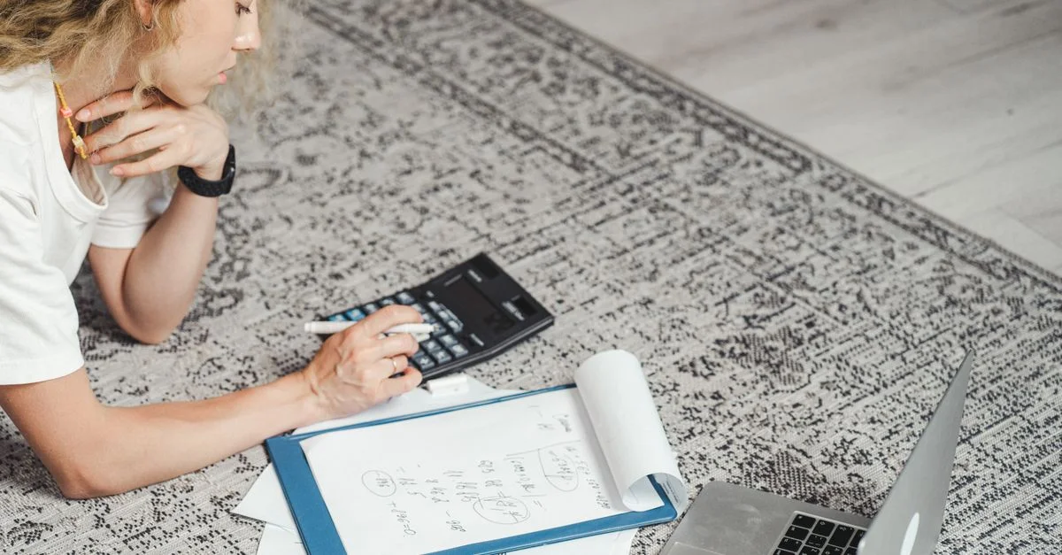 Une femme calcule ses finances à la maison en utilisant un ordinateur portable, une calculatrice et des documents sur un tapis.