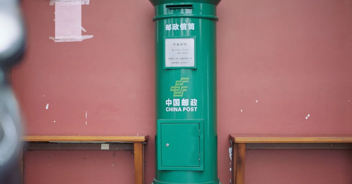 Dans un cadre urbain typique, une boîte aux lettres de China Post se dresse contre un mur rouge, encadrée de bancs.