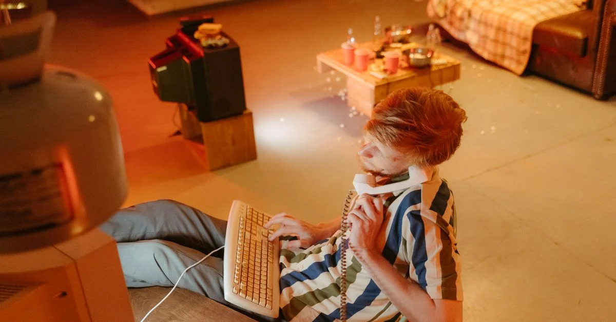 Un homme plongé dans la technologie des années 80, utilisant un ordinateur et un téléphone dans une pièce au style rétro.