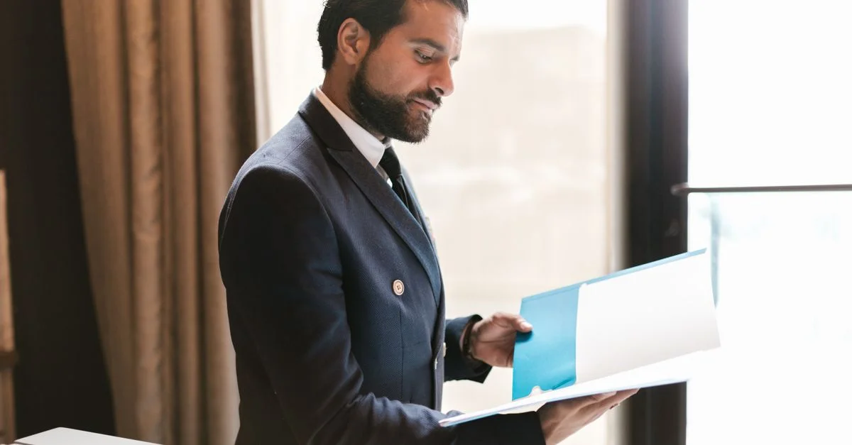 Homme d'affaires barbu tenant un dossier près d'une fenêtre, vêtu d'un costume noir dans un bureau.