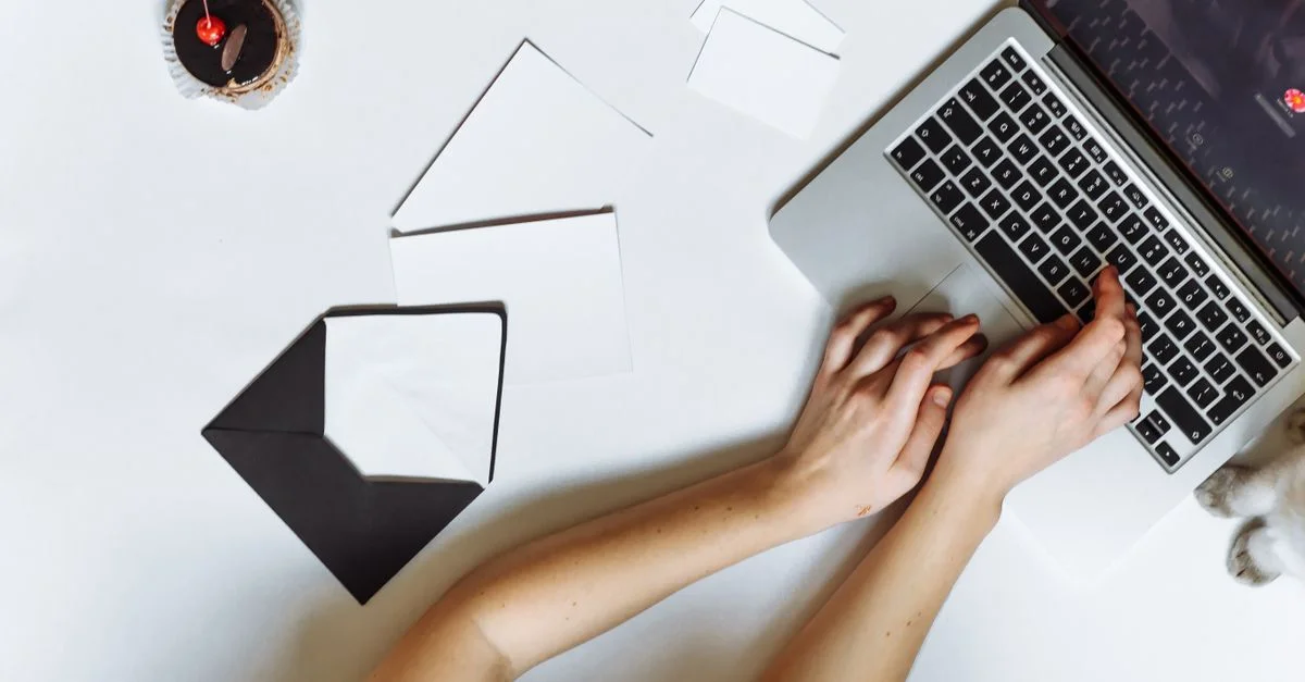 Vue de dessus d'une personne tapant sur un ordinateur portable avec des enveloppes et un cupcake sur un bureau blanc.
