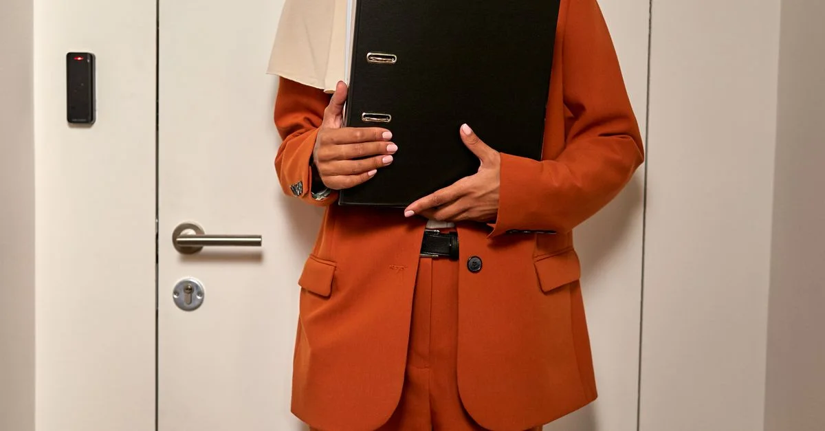 Gros plan sur une femme en costume orange tenant des documents, debout dans un environnement de bureau.