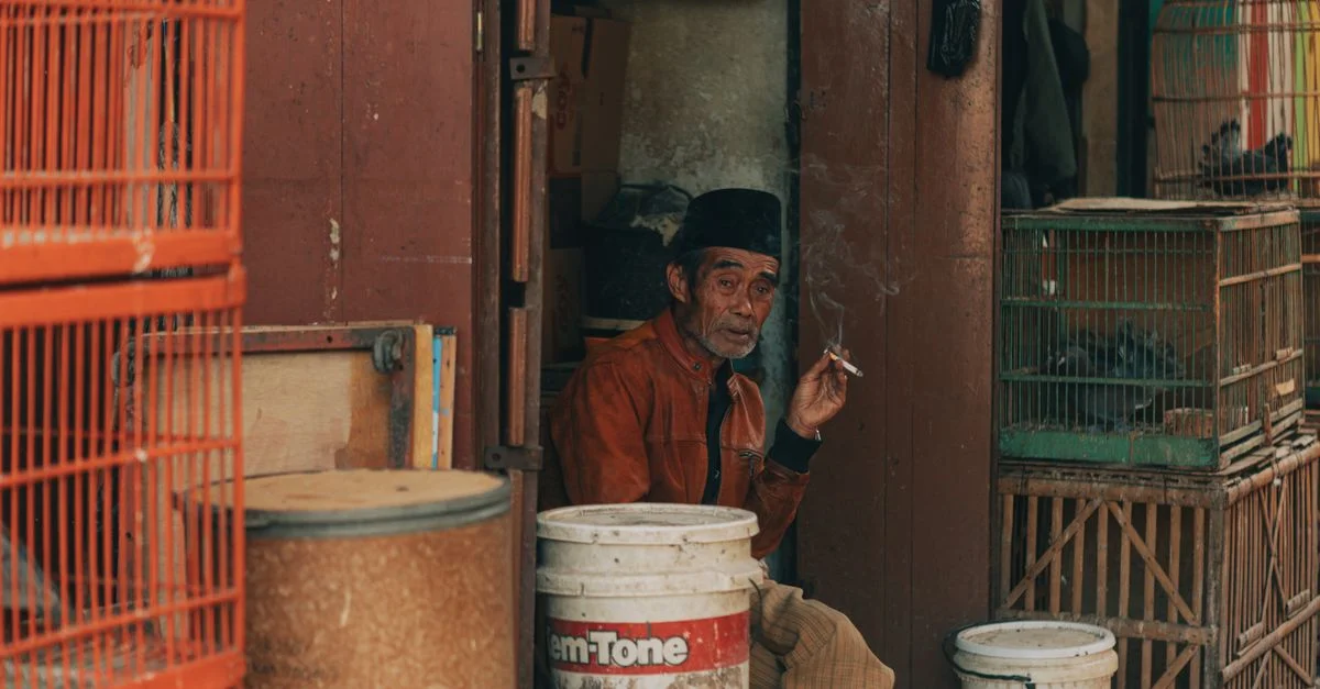 À Java Ouest, en Indonésie, un homme fume une cigarette devant une boutique rustique entourée de cages à oiseaux colorées.