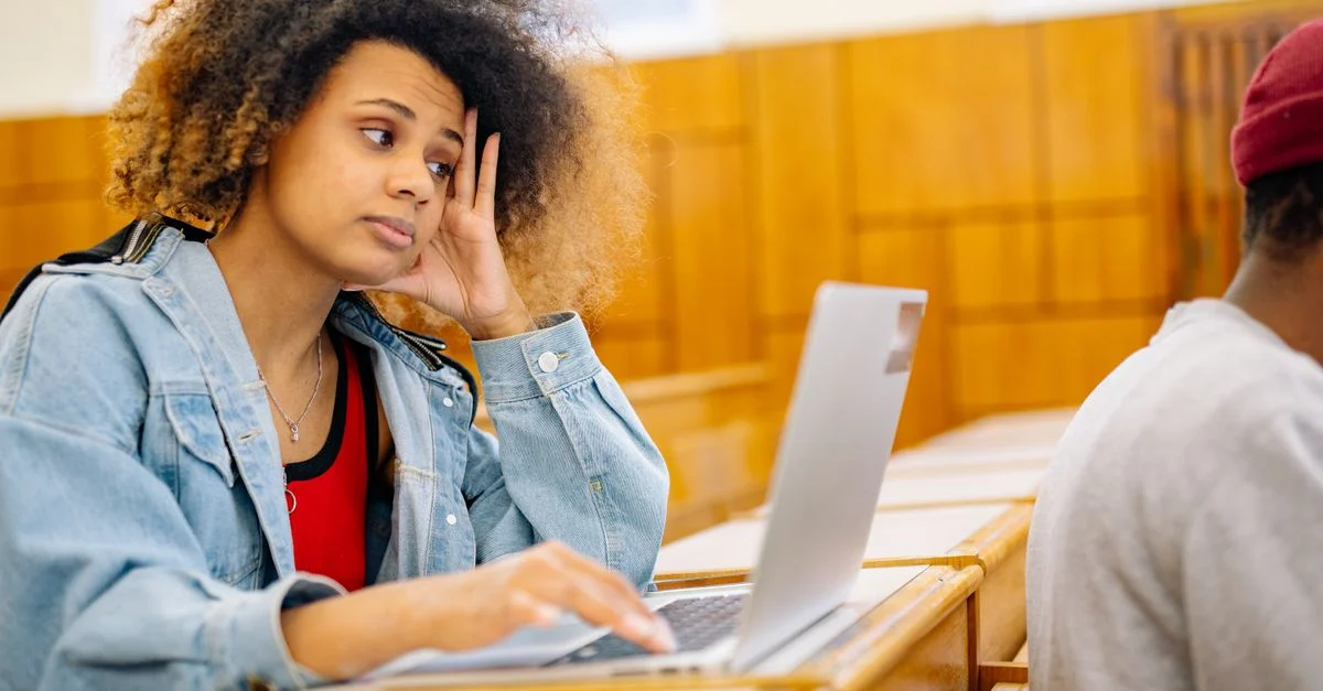 Une jeune femme est assise dans une salle de classe, concentrée sur son ordinateur portable, illustrant une session d'étude.