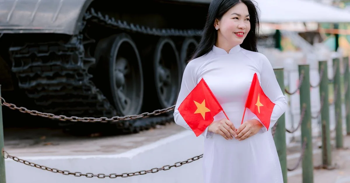 Une femme vietnamienne en tenue traditionnelle tient des drapeaux nationaux près d'un char historique à l'extérieur.