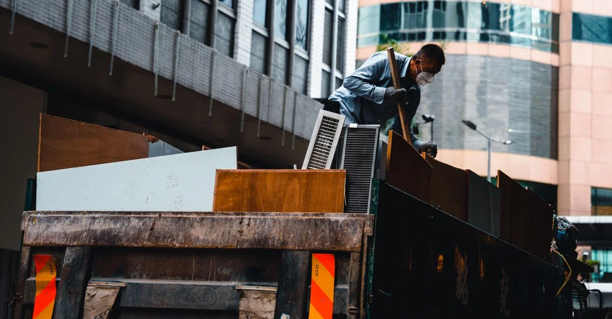 Un homme portant un masque gère des débris de construction dans un environnement urbain depuis un grand camion.