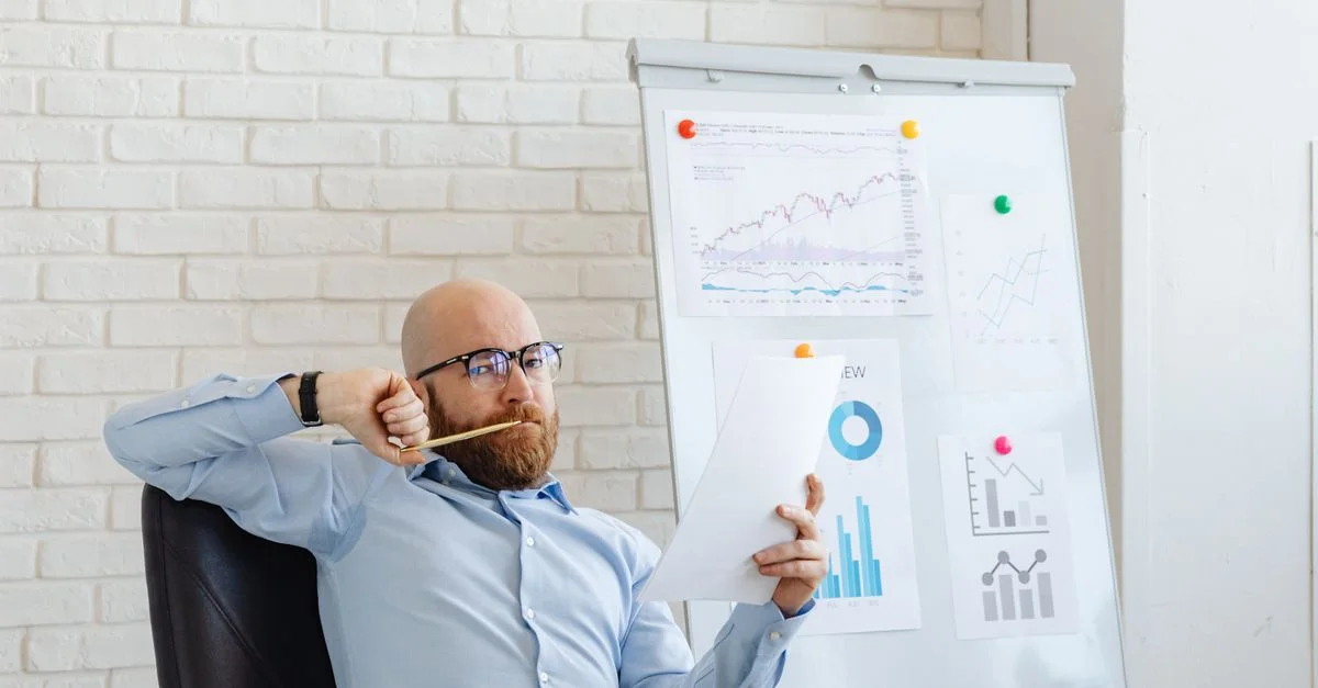 Un homme d'affaires portant des lunettes examine des graphiques financiers, pensant profondément devant un tableau blanc avec des graphiques.