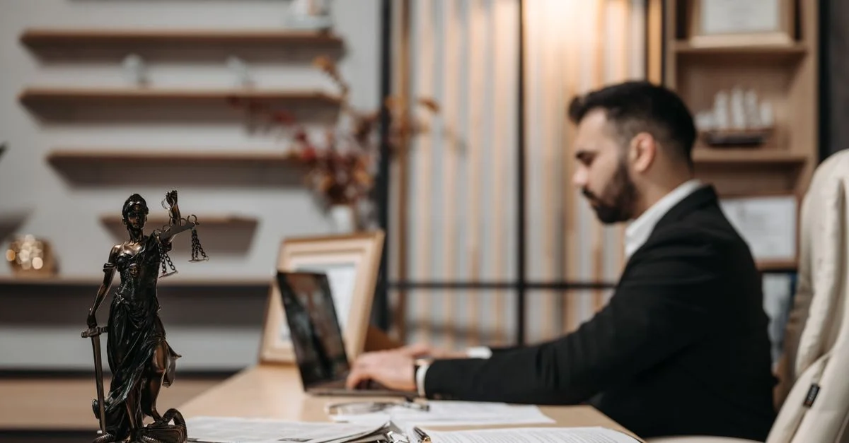 Avocat travaillant avec diligence sur un ordinateur portable dans un bureau moderne, statue de la Justice présente.