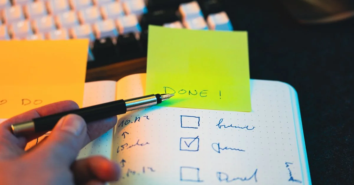 Gros plan sur une main tenant un stylo au-dessus d'une liste de contrôle dans un carnet, avec des notes autocollantes portant la mention « TERMINÉ ! »