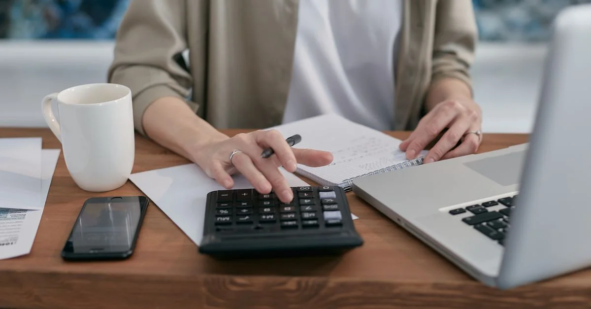 Une personne utilisant une calculatrice et un ordinateur portable tout en prenant des notes à un bureau en bois.