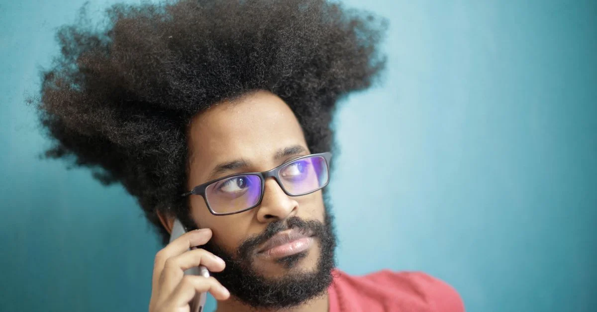 Jeune homme aux cheveux afros en train de passer un appel téléphonique à l'intérieur sur fond bleu.