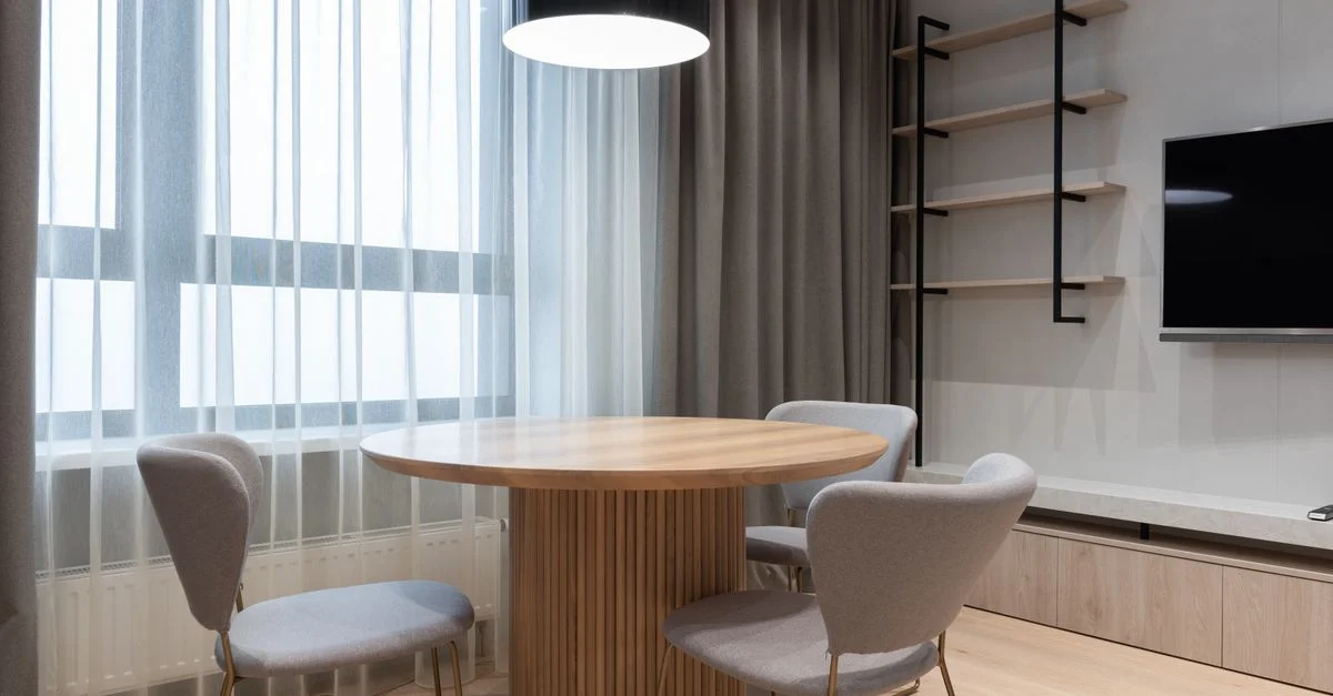 Salon lumineux et minimaliste avec une table ronde en bois, des chaises modernes et une télévision murale.