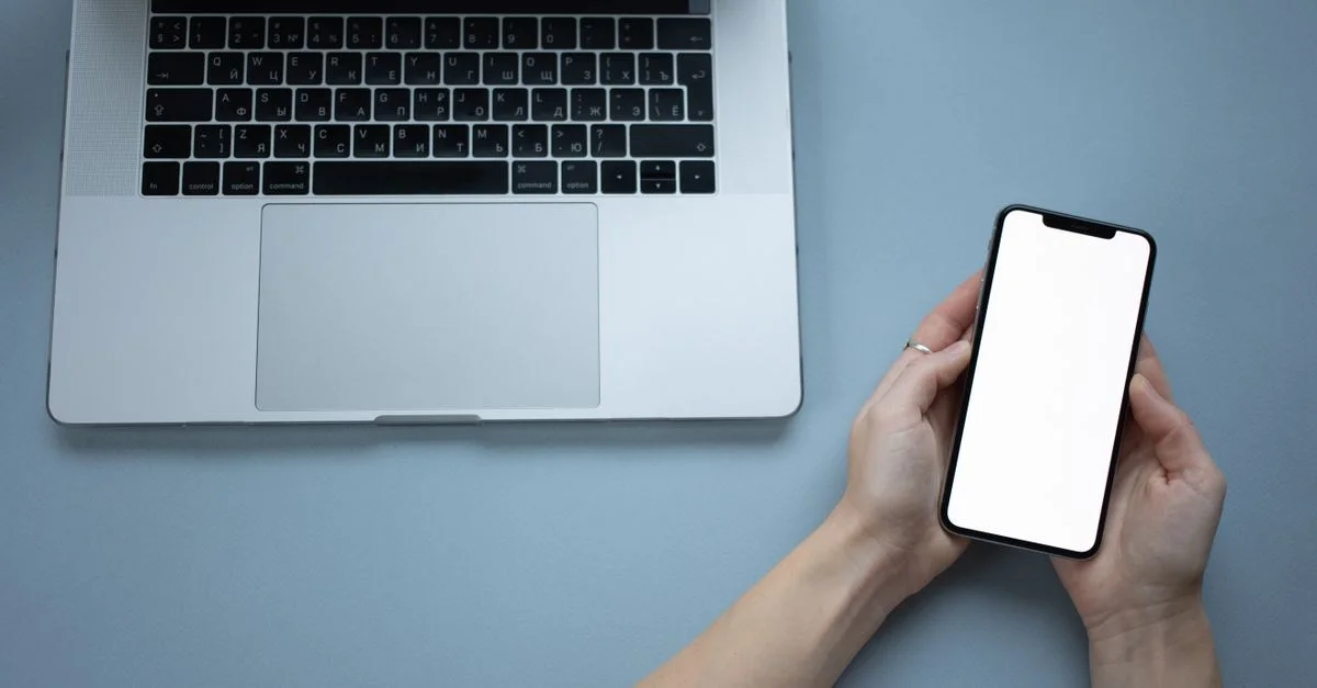 Des mains tenant un smartphone avec un écran vide à côté d'un ordinateur portable, idéal pour des maquettes.