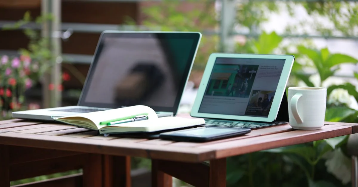 Une scène de bureau extérieur confortable avec un ordinateur portable, une tablette et une tasse sur une table en bois.
