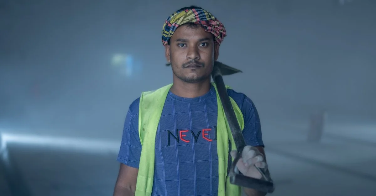 Un ouvrier du bâtiment, tenant une pelle, portant un gilet de sécurité et un foulard, dans un espace industriel.