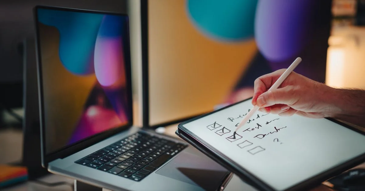 Une main écrivant sur une tablette avec un stylet dans un bureau contemporain avec des ordinateurs portables et des écrans numériques.