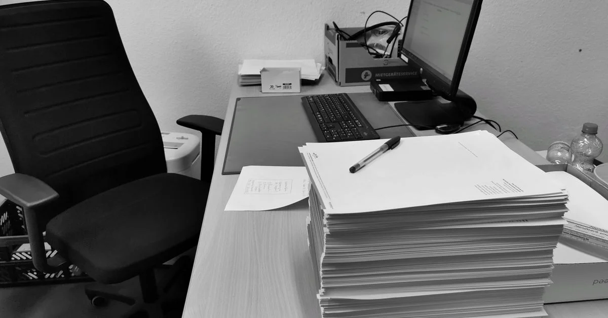 Image en noir et blanc d'un bureau encombré avec un ordinateur, une chaise et des papiers.