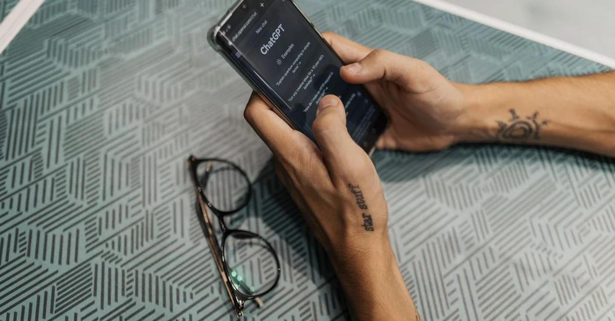 Gros plan sur une personne tenant un smartphone montrant l'application ChatGPT à côté de lunettes.