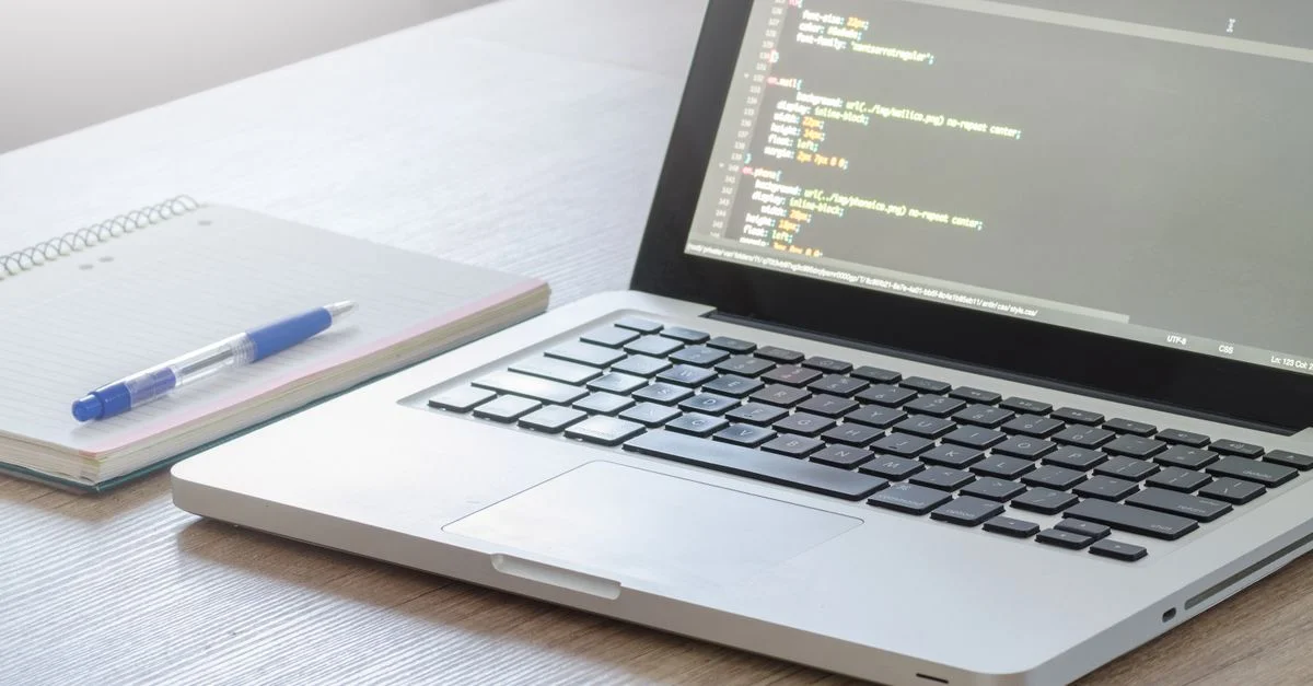 Ordinateur portable avec éditeur de code ouvert, carnet et stylo sur un bureau en bois, parfait pour des thèmes technologiques et de travail.