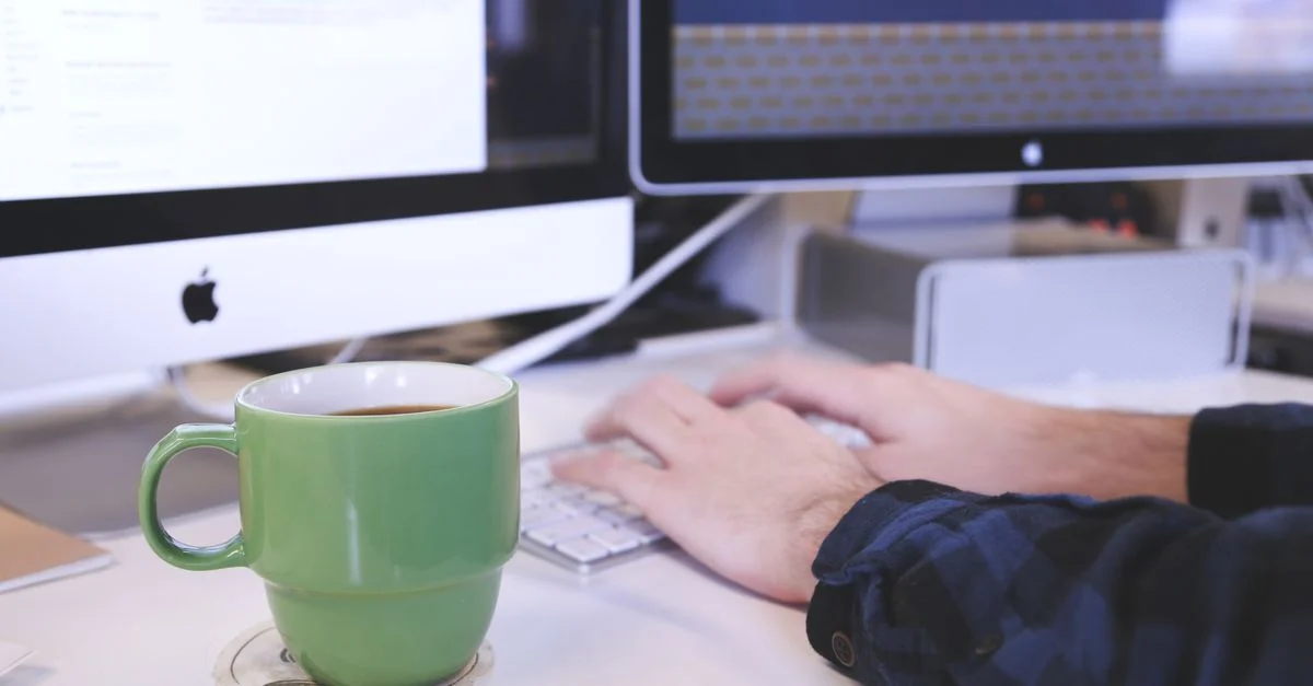 Personne tapant à un bureau avec du café et des écrans d'ordinateur. Parfait pour des thèmes d'entreprise ou de technologie.