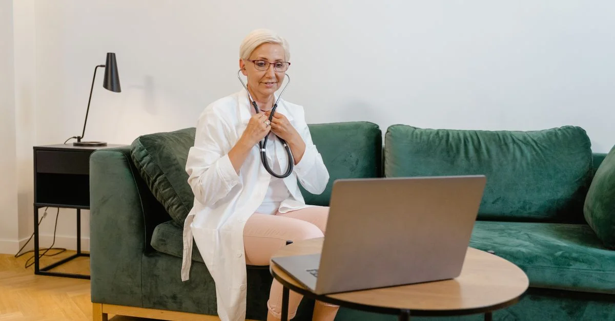Médecin femme en blouse blanche effectuant une consultation en ligne depuis chez elle à l'aide d'un ordinateur portable.