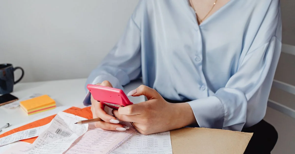 Une femme utilisant une calculatrice rose entourée de factures et de reçus sur un bureau.