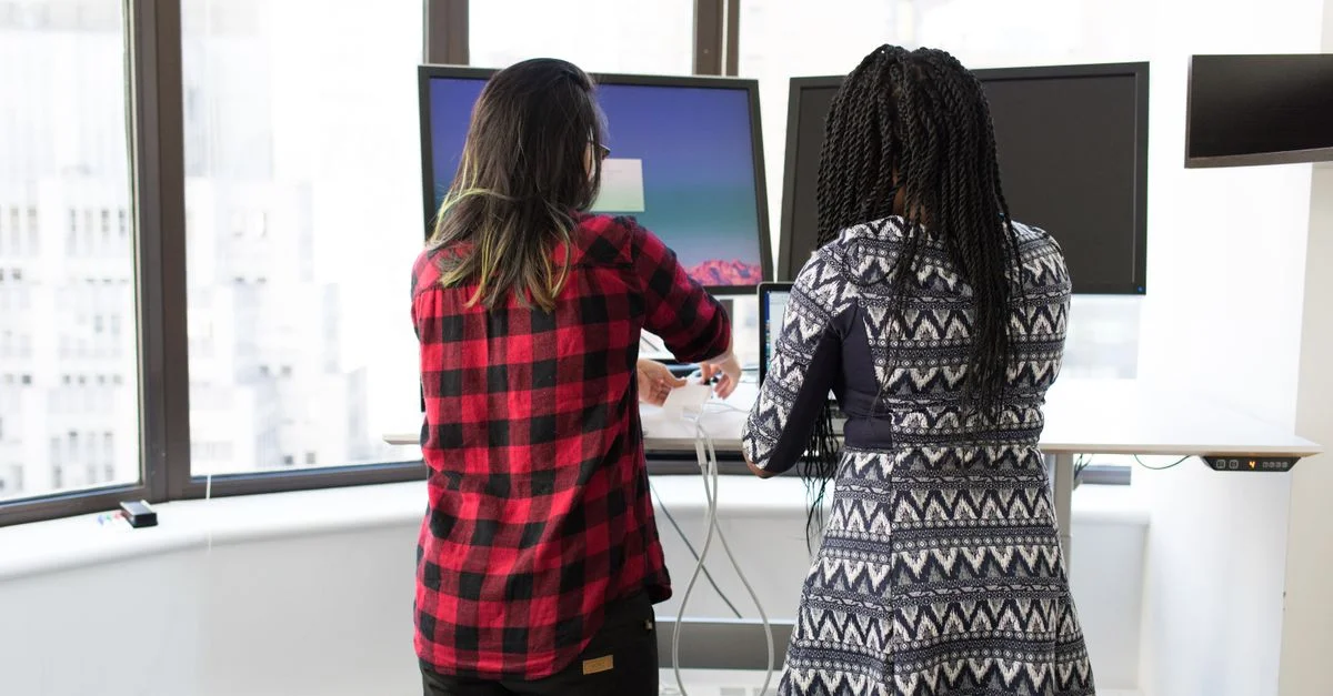 Deux femmes travaillant ensemble à des bureaux debout dans un bureau moderne et lumineux. Accent sur la technologie et la collaboration.