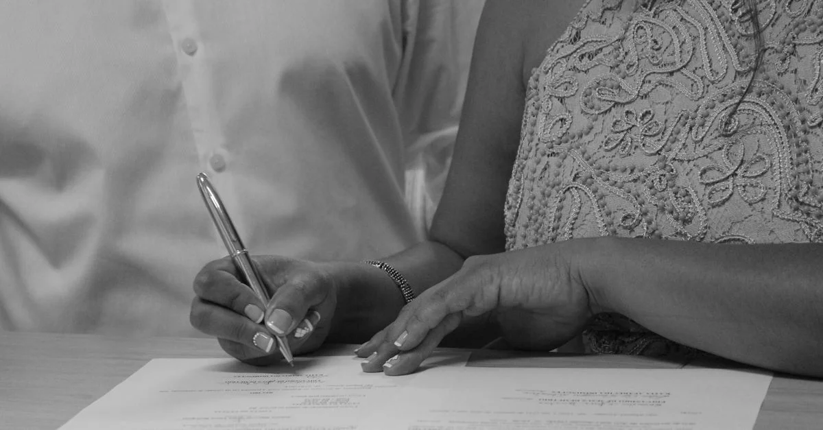 Image en noir et blanc d'un couple signant des documents, mettant l'accent sur leurs mains et leur stylo.